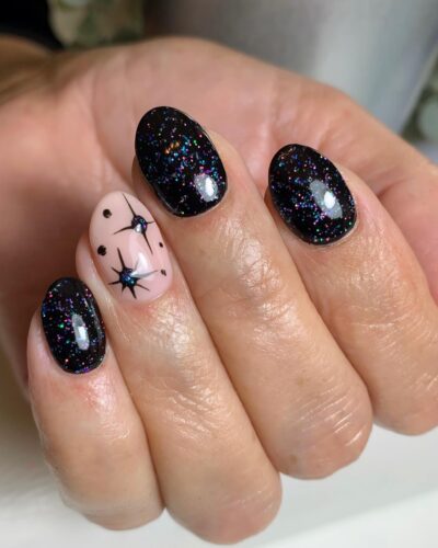 Close-up of hands with black sparkly nails resembling a starry night sky and a single nude nail with delicate black star designs.