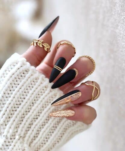 Long stiletto nails alternately designed with matte black, glossy black, and sparkling gold, adorned with elegant gold band accents.