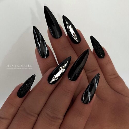 Stiletto nails with a glossy black finish, some adorned with silver glitter and jewels, embodying a gothic glam aesthetic.