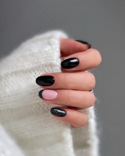 A hand with a mix of glossy black, glittery black, and one nude accent nail, showcasing a variety of textures.