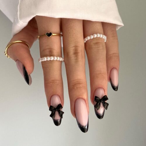 Stiletto nails with black tips and transparent bases, adorned with cute black bows at the transition point.