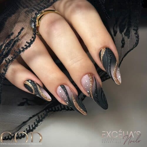 Hands draped in a lace garment displaying long almond-shaped nails with a black base, gold glitter accents, and semi-transparent panels.