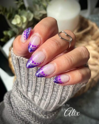 Almond-shaped nails with a speckled purple and glitter design on a cozy grey knitted sweater background.