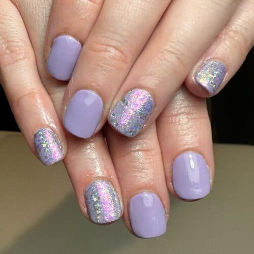 Short rounded nails with a lavender base and a chunky glitter gradient against a neutral background.