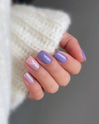 Rounded nails with alternating lilac and silver glitter polish on a fluffy white sweater background.