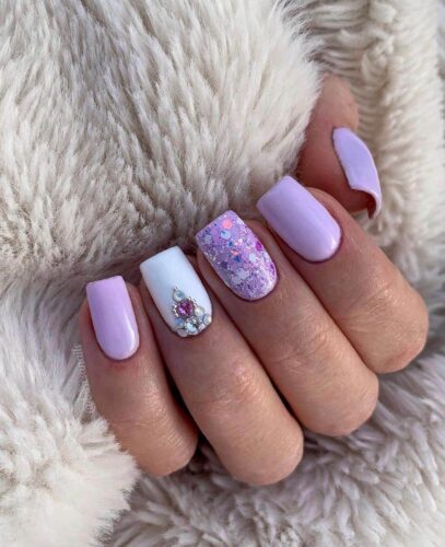 A mix of pastel purple nails, one with glitter and another with rhinestone embellishments, against a plush furry background.