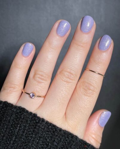 Rounded nails painted with pastel purple polish and a subtle sprinkle of glitter against a dark sweater background.