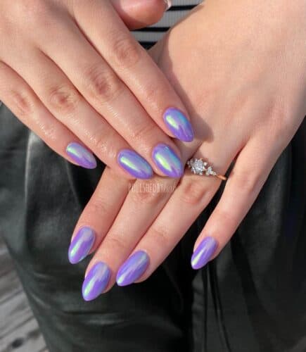 Almond-shaped nails with an opalescent purple finish that reflects a range of colors, held against a black leather backdrop.