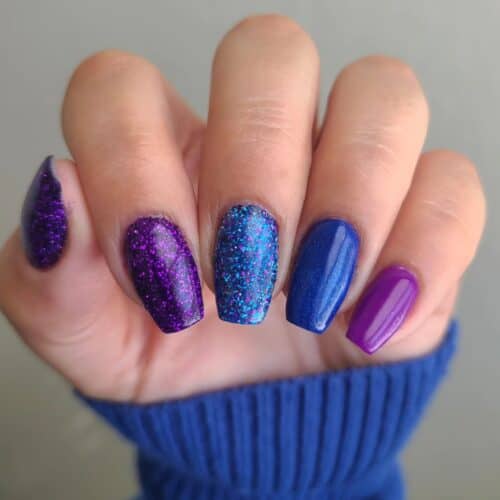 A mixture of purple and blue nails with different textures and densities of glitter against a soft blue sweater.