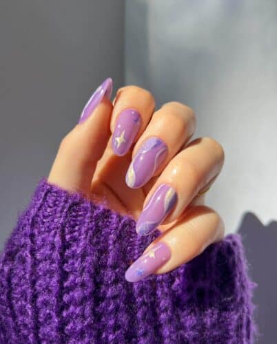 Lavender nails with gold star accents and a sprinkle of glitter, cradled in a cozy purple knitted sweater.