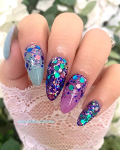 Festive nails with a bold mix of purple and green glitter against a delicate white lace background.