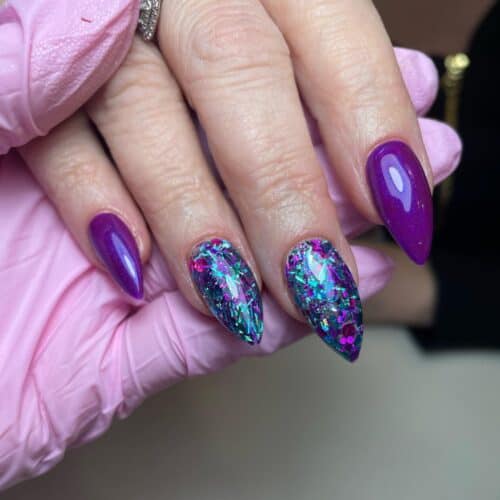 Stiletto nails showcasing a vibrant purple and iridescent teal glitter design against a pink glove.