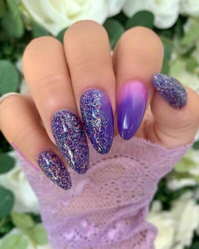 Long nails covered in purple glitter with one nail featuring a purple ombre effect, held against a background of white roses.
