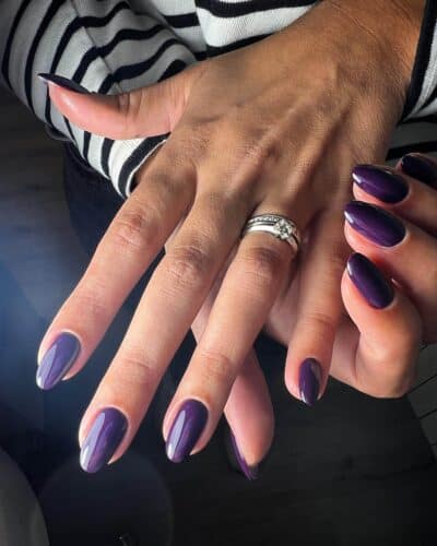Almond-shaped nails polished in a glossy, deep royal purple shade against a striped and dark background.