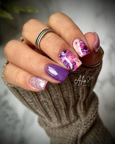 A mix of glossy purple nails, two with a floral design and another with purple glitter, held against a cozy knit sweater. another with purple glitter, held against a cozy knit sweater.