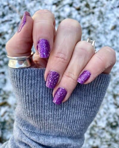 Short, rounded nails covered in frosty purple glitter against a snowy backdrop.