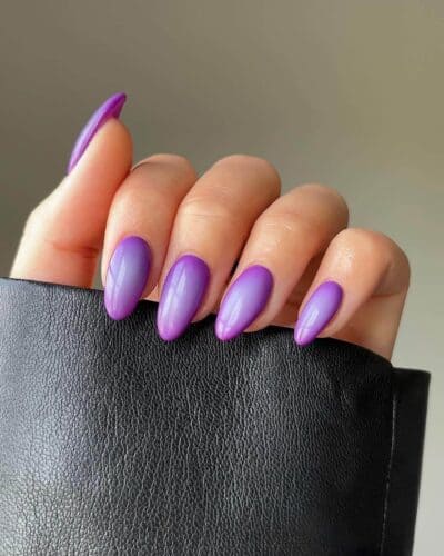 Long, stiletto nails with a smooth gradient from clear to purple, presented against a leather backdrop.