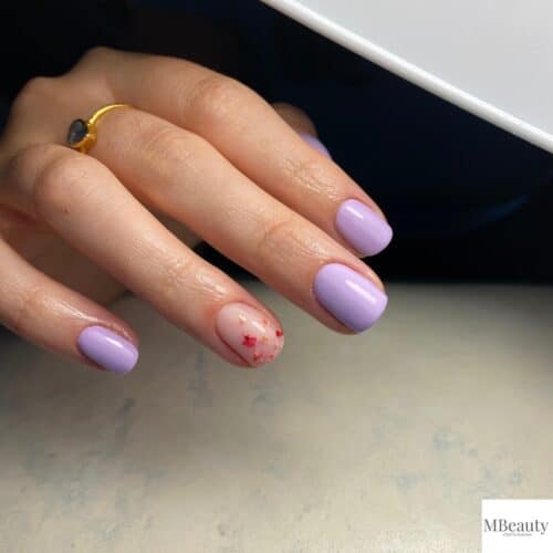 Short, rounded nails painted in a soft lavender shade, with one nail featuring tiny red floral accents.