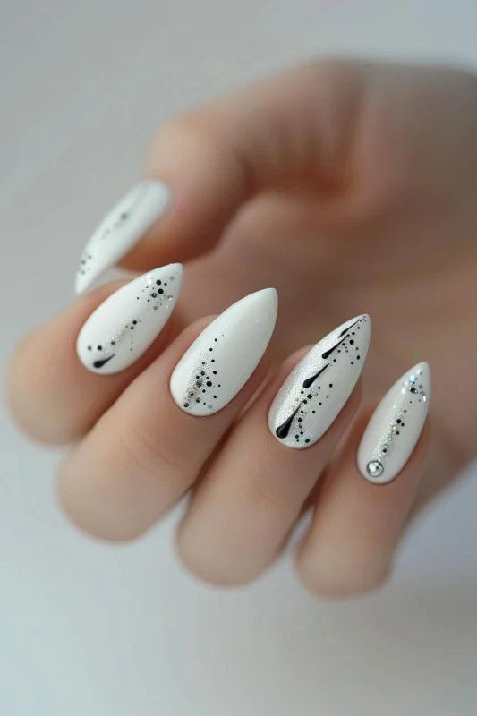 White almond-shaped nails with black splatter design and gemstone accents.
