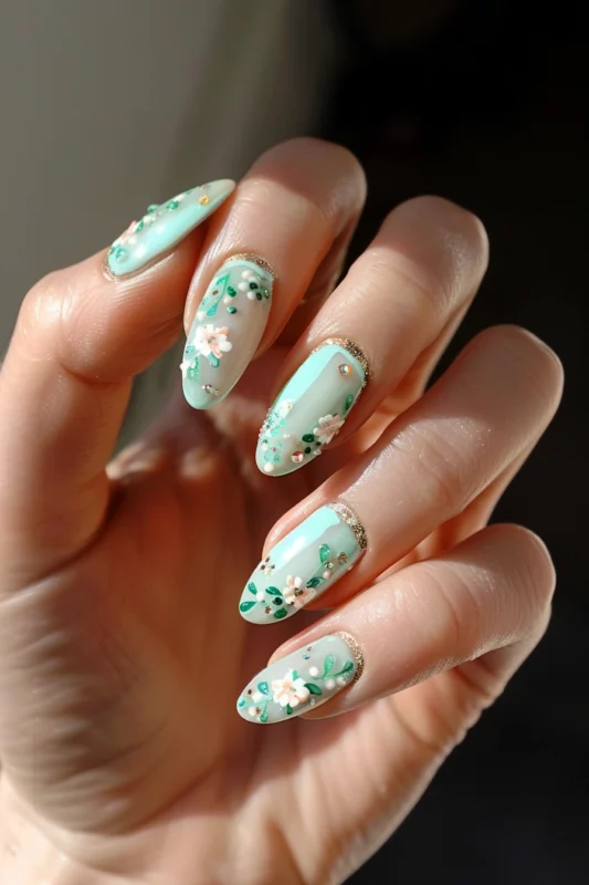 Green nails with a white and pink floral design and a glittery gold stripe at the base of the nail.