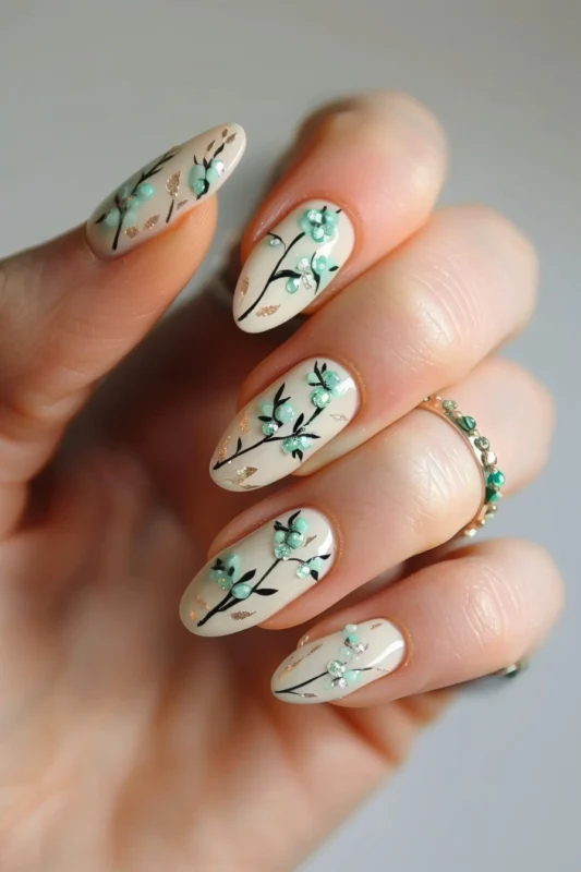 A hand with almond-shaped nails, decorated with spring-themed nail art featuring green accents gem and gold details.