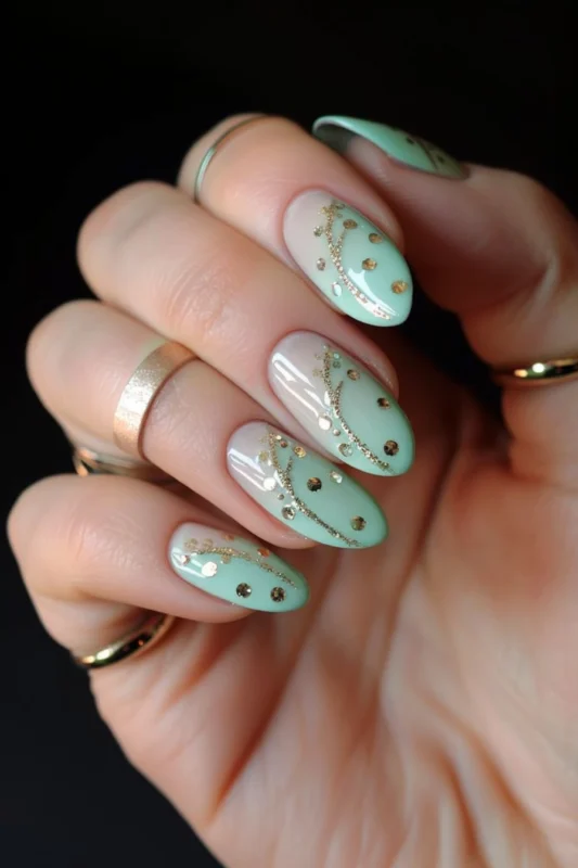 A hand with almond-shaped nails in light green ombre embellished with gold swirls and gem accents.