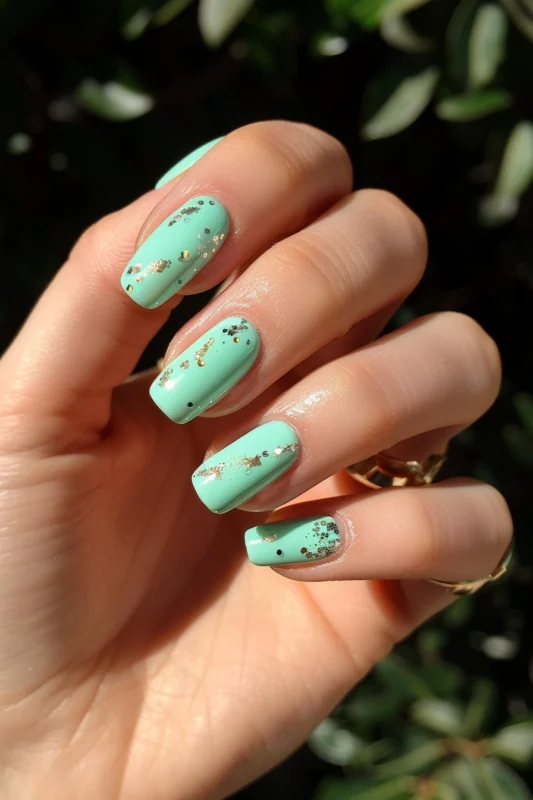 Short, square nails in a light green nail polish decorated with gold speckles.