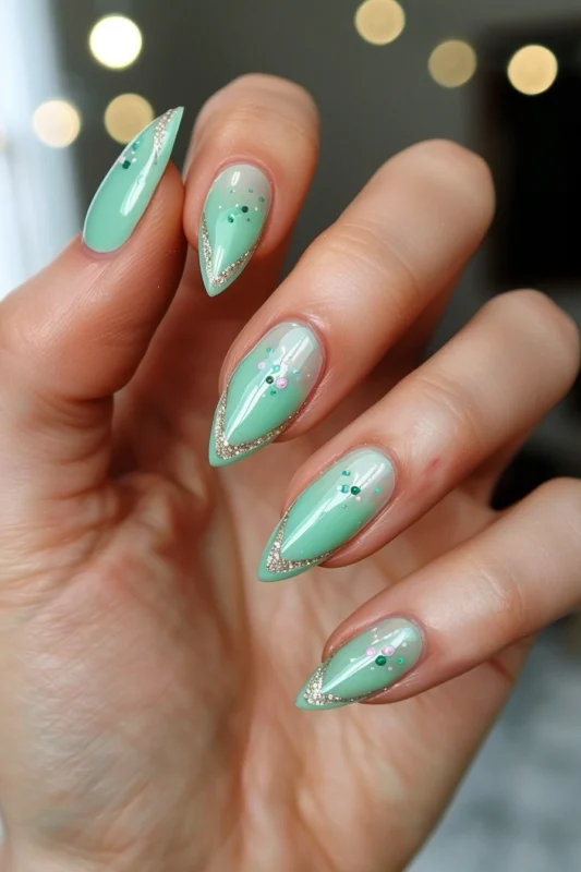 A hand with long pointy nails featuring a light green ombre base and a golden glitter French tip.