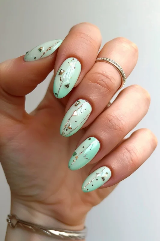 Almond-shaped nails with a mint green base and geometric gold studs.
