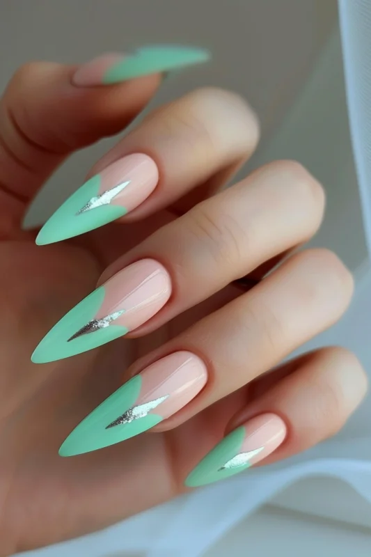 A hand with long stiletto nails featuring pastel minty green French tips with a silver glitter accent.