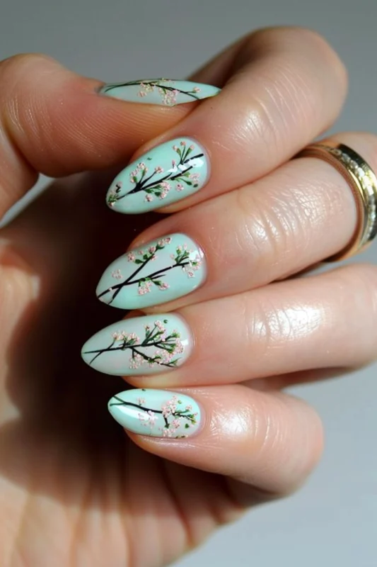 A hand with long, oval nails painted with cherry blossom branches on a pastel green background.
