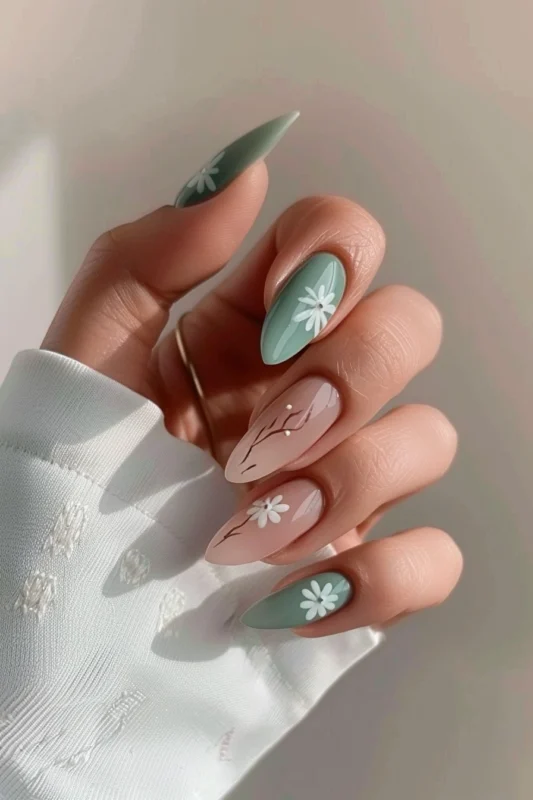 A hand with long nails painted in alternating sage green and blush, adorned with white floral designs.