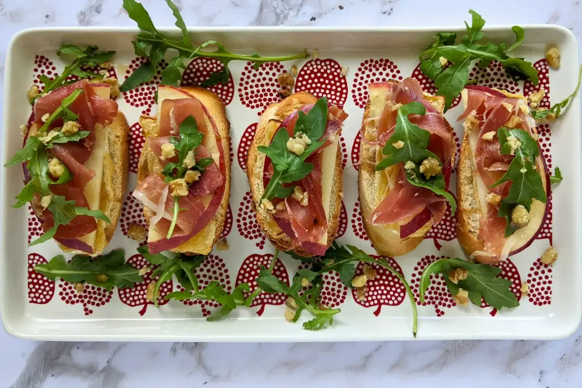 Crostini appetizers with apple, Brie cheese, and serrano ham topped with arugula, crushed walnuts and maple syrup.