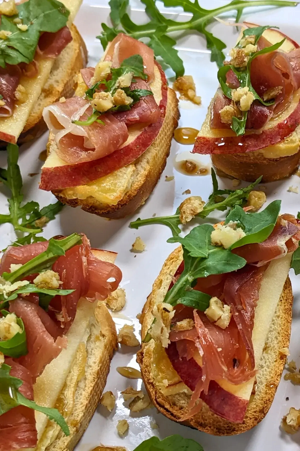 Crostini appetizers with apple, Brie cheese, and serrano ham topped with arugula, crushed walnuts and maple syrup.