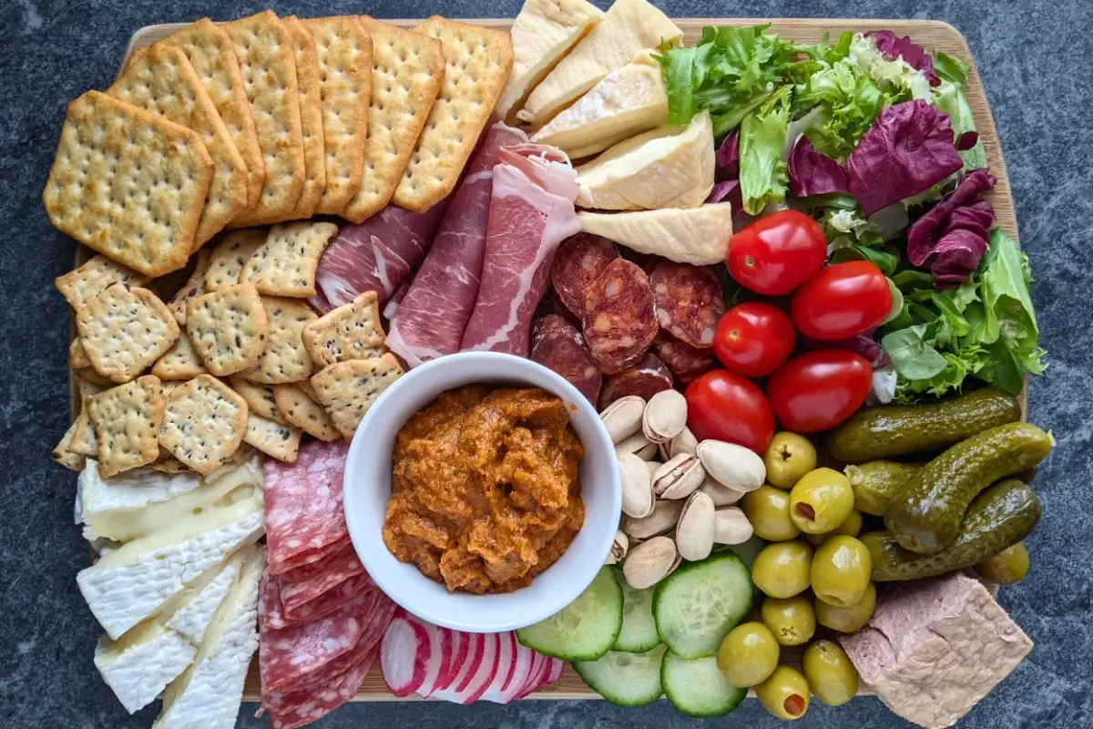 A healthier charcuterie appetizer board featuring fresh vegetables, soft cheeses, sliced meats, and pickled bites on a rustic board.