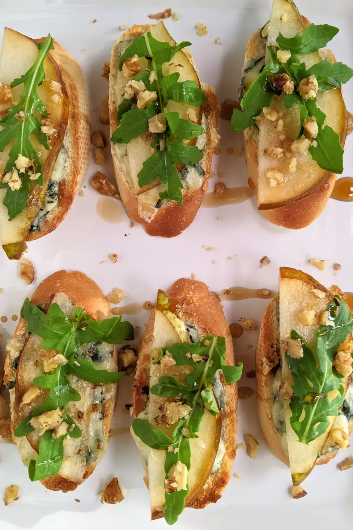 Crostini appetizers with pear and blue cheese, topped with arugula, crushed walnuts, and maple syrup.