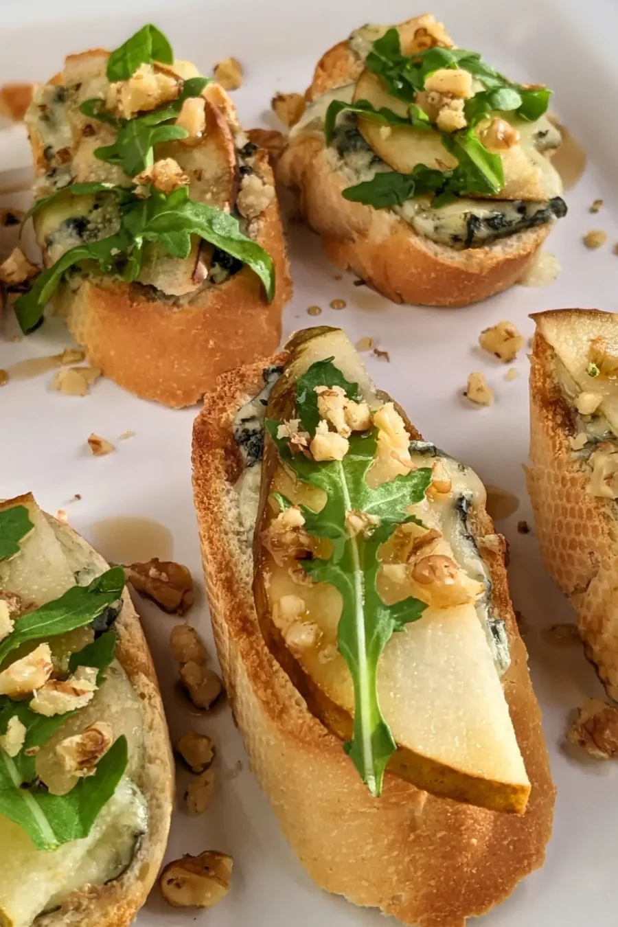 Crostini appetizers with pear and blue cheese, topped with arugula, crushed walnuts, and maple syrup.