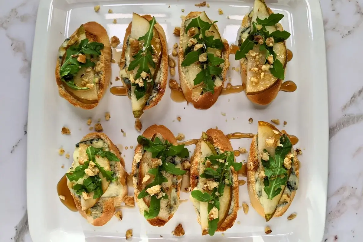Crostini appetizers with pear and blue cheese, topped with arugula, crushed walnuts, and maple syrup.