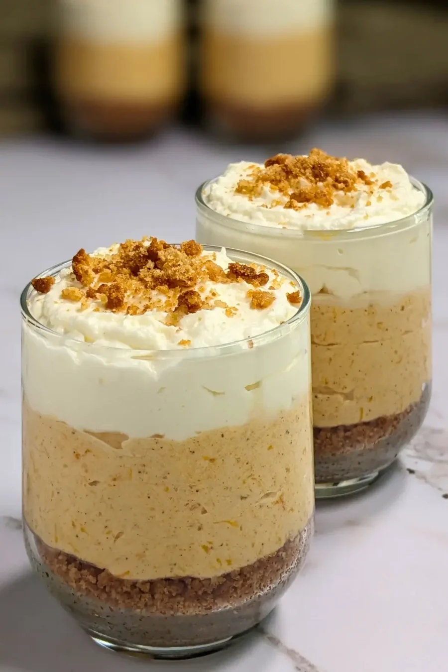 Pumpkin cheesecake mousse cups layered with a cookie and butter base; cream cheese, pumpkin, and whipped cream mousse; and topped with more whipped cream.