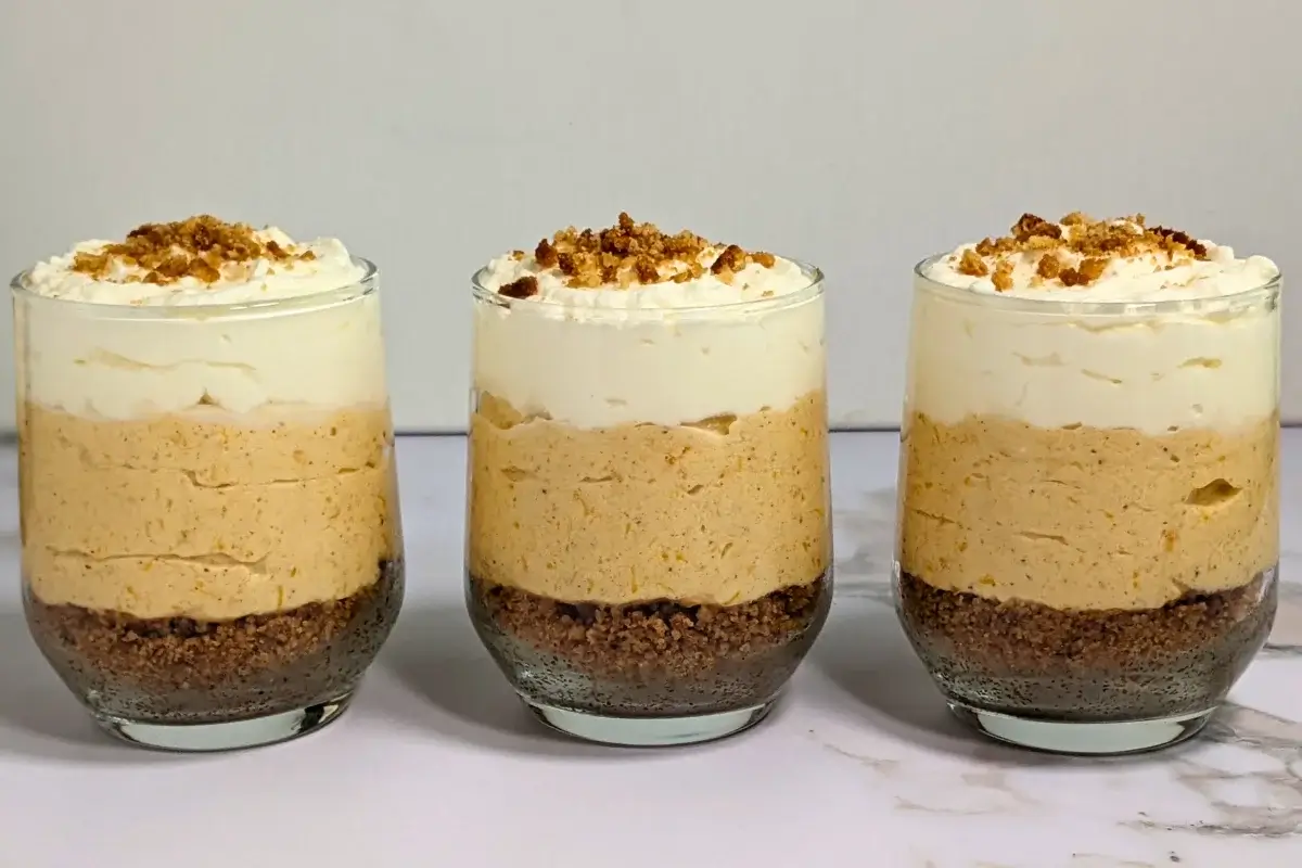 Pumpkin cheesecake mousse cups layered with a cookie and butter base; cream cheese, pumpkin, and whipped cream mousse; and topped with more whipped cream.