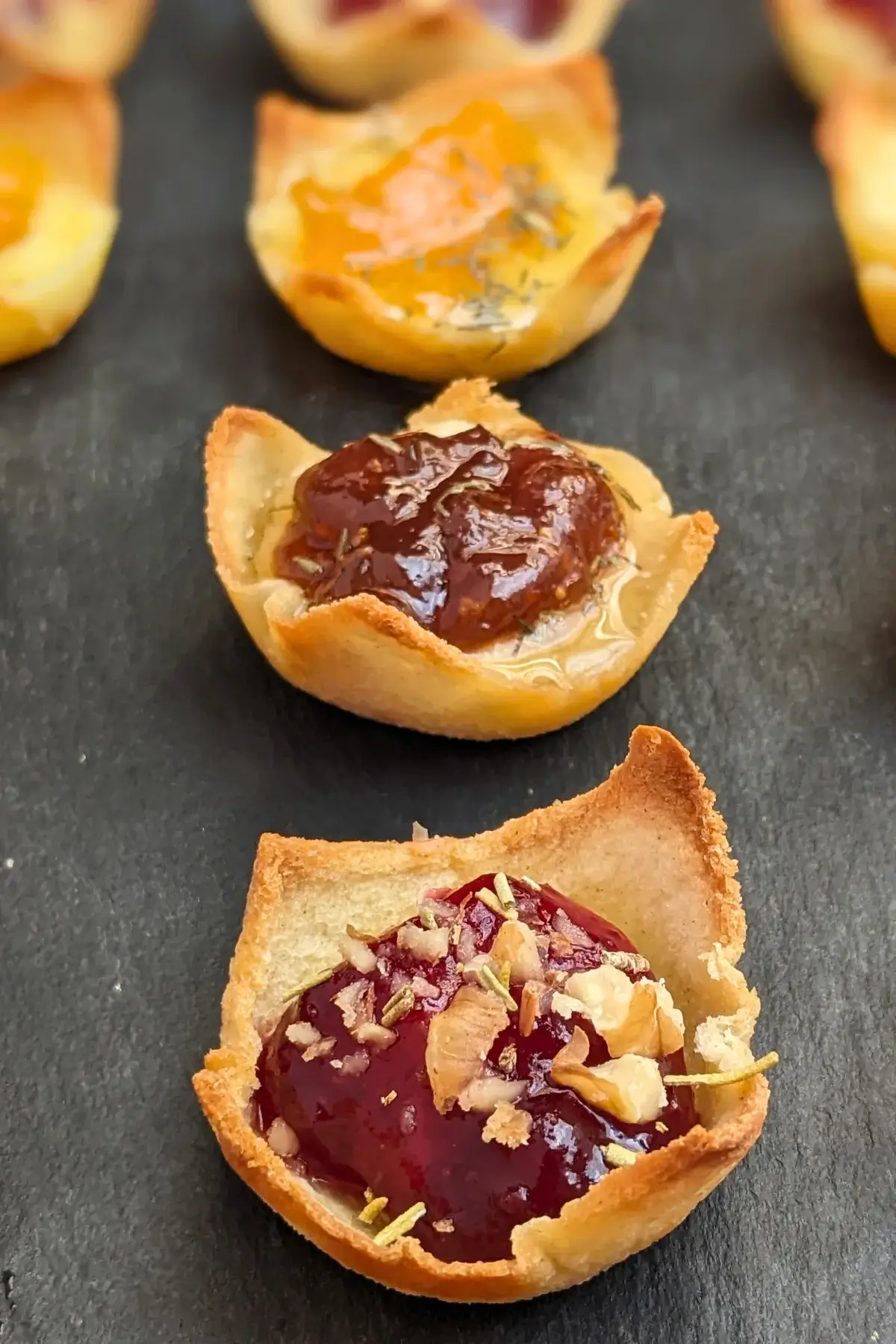 Trio of cheese and jam appetizers in mini toast cups - mini bread toast cups filled with Brie cheese and cranberry jam, Parmesan cheese and fig jam, and cheddar cheese and apricot jam, topped with crushed walnuts and a honey-dried thyme drizzle.