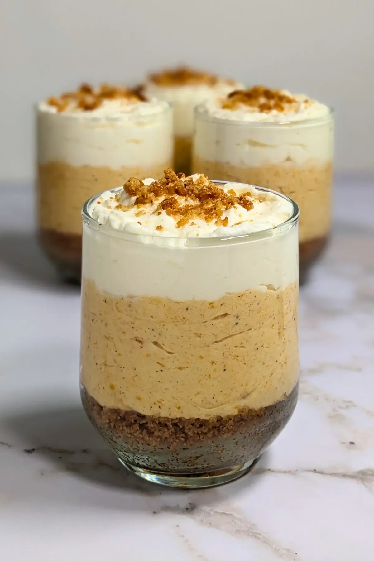 Pumpkin cheesecake mousse cups layered with a cookie and butter base; cream cheese, pumpkin, and whipped cream mousse; and topped with more whipped cream.