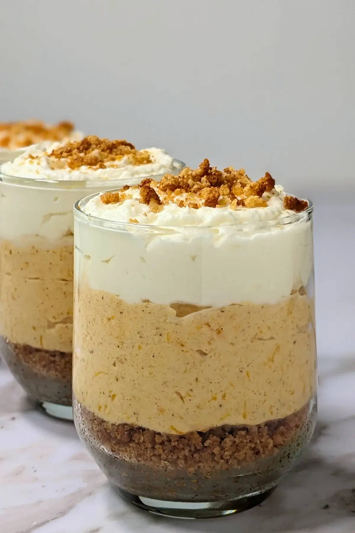 Pumpkin cheesecake mousse cups layered with a cookie and butter base; cream cheese, pumpkin, and whipped cream mousse; and topped with more whipped cream.