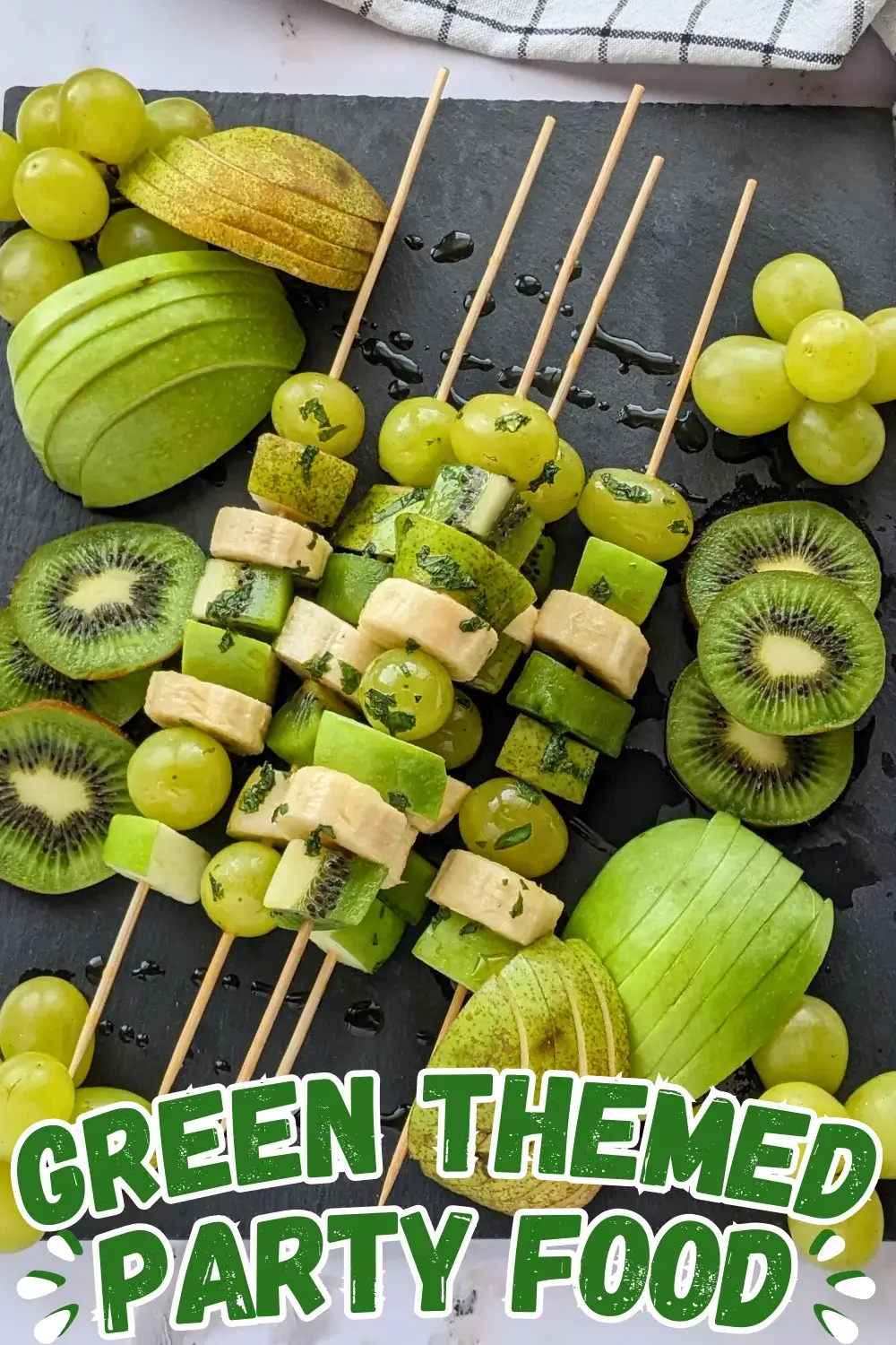 Green Fruit Tray - colorful green-themed fruit tray with green fruit kabobs featuring fruits like kiwi, green grapes, green apple, green pear, and bananas that are drizzled with a fresh mint, lime juice, and maple sryup sauce, plus slices of green apple, green pear, kiwis, and little piles of green grapes.