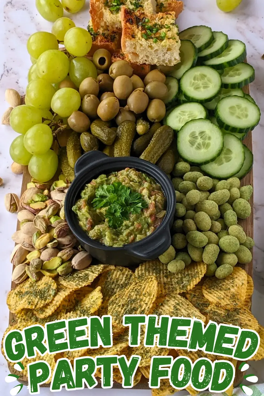 Green Snack Board - Green themed snack board and party tray with homemade garlic bread and guacamole dip, dried chives covered potato chips, fresh cucumbers, mini pickles, olives, pistachios, crunchy wasabi coated peanuts, and green grapes.