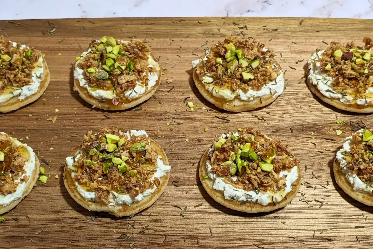 Mini Pancake Bites With Dried Onions - savory mini pancake canapes topped with a creamy herb-and-garlic cream cheese spread, crispy dried onions, maple syrup, and crushed pistachios for garnish.