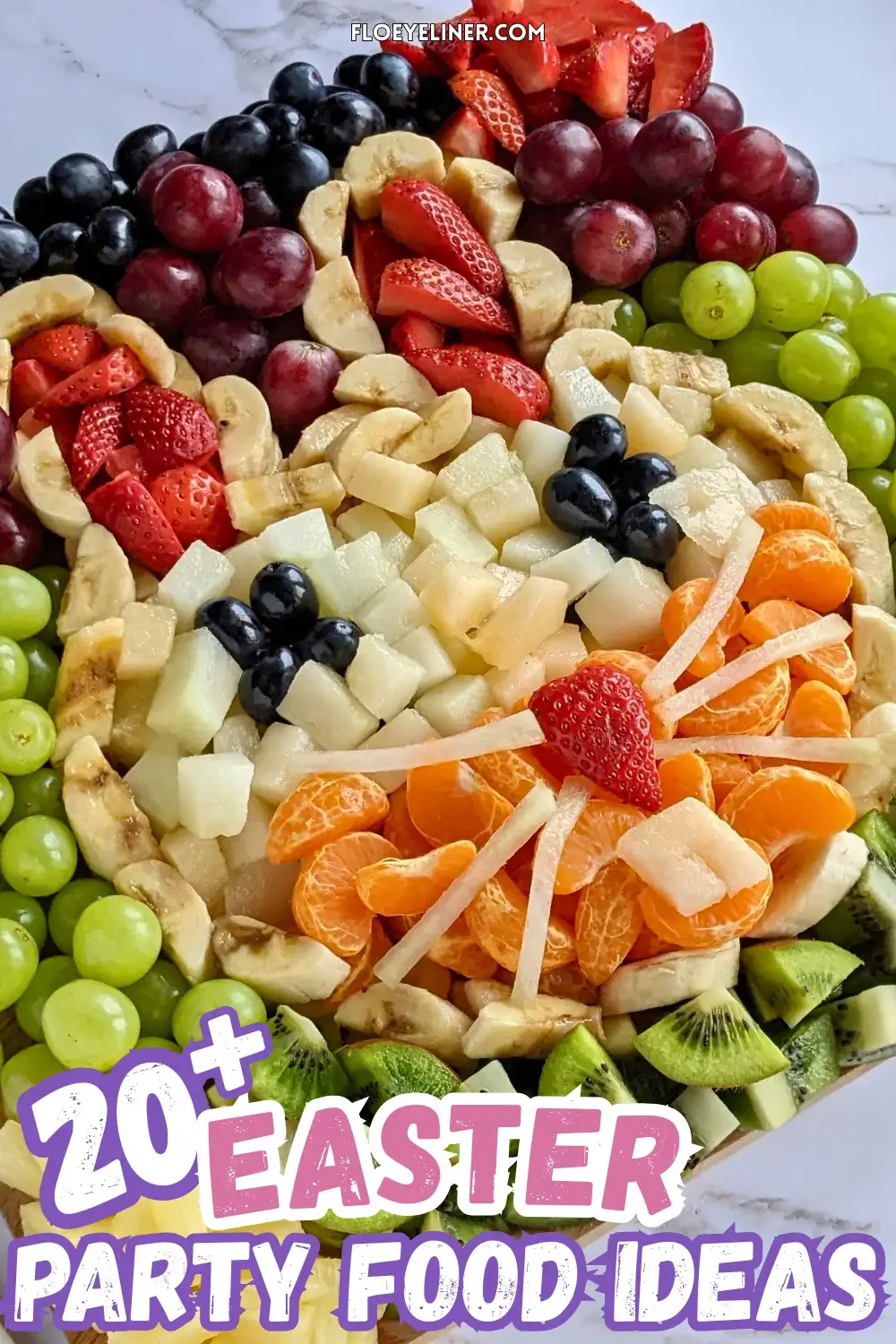 Easter Bunny Fruit Tray - Easter fruit tray in an Easter bunny theme, with the bunny head shaped with banana pieces, filled with sliced strawberries for ears, blue grapes for eyes, and pieces of yellow melon and mandarin segments, surrounded by colorful fruits like dark red, blue, and green grapes; canned pineapple pieces; kiwi pieces; and sliced srawberries.