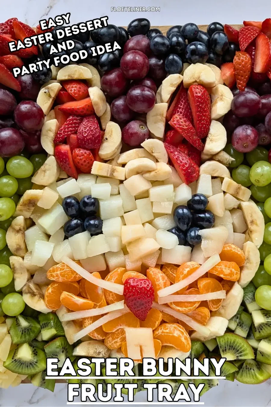 Easter Bunny Fruit Tray - Easter fruit tray in an Easter bunny theme, with the bunny head shaped with banana pieces, filled with sliced strawberries for ears, blue grapes for eyes, and pieces of yellow melon and mandarin segments, surrounded by colorful fruits like dark red, blue, and green grapes; canned pineapple pieces; kiwi pieces; and sliced srawberries.