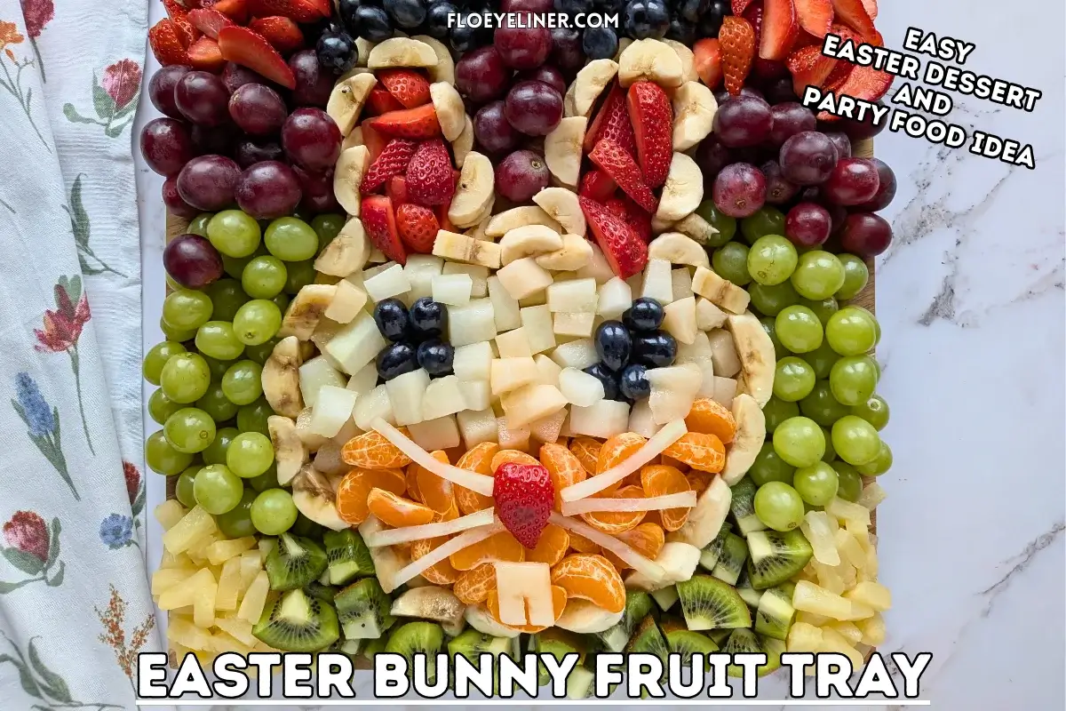 Easter Bunny Fruit Tray - Easter fruit tray in an Easter bunny theme, with the bunny head shaped with banana pieces, filled with sliced strawberries for ears, blue grapes for eyes, and pieces of yellow melon and mandarin segments, surrounded by colorful fruits like dark red, blue, and green grapes; canned pineapple pieces; kiwi pieces; and sliced srawberries.